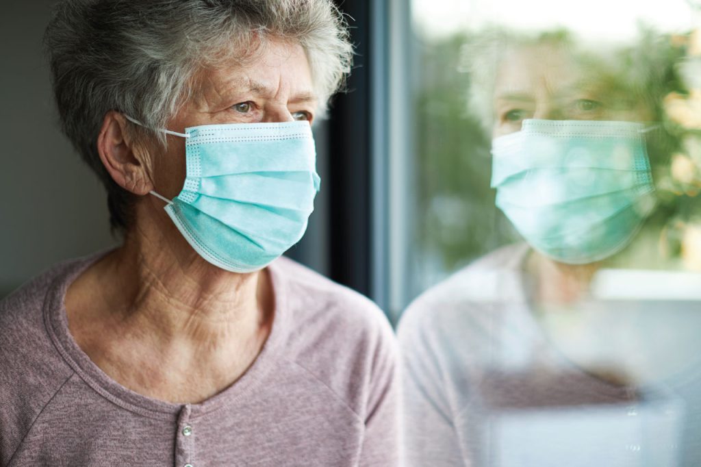 An older woman wearing a sanitation mask looking out a window